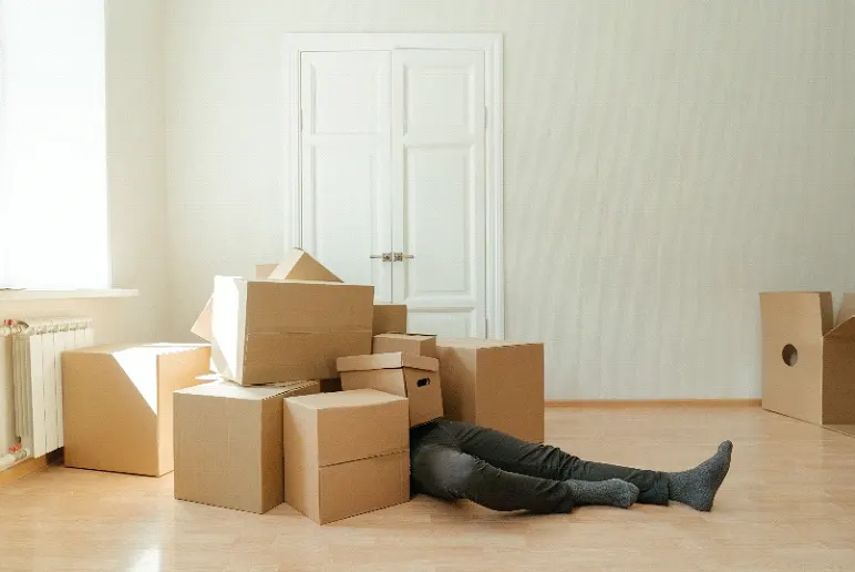 Image of moving boxes on top of a person lying on the floor with only their legs visible.